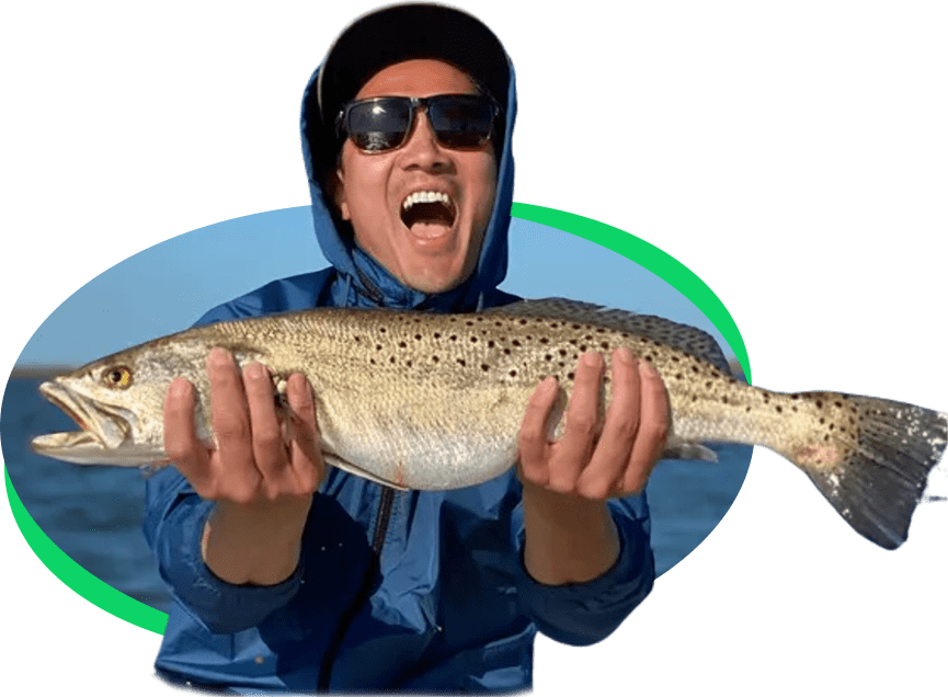 A man in sunglasses and a cap excitedly holds a large trout in front of a body of water.