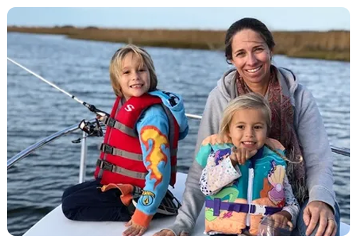 A woman smiles on a boat with two young children in life jackets, all looking at the camera, with water and the horizon in the background.